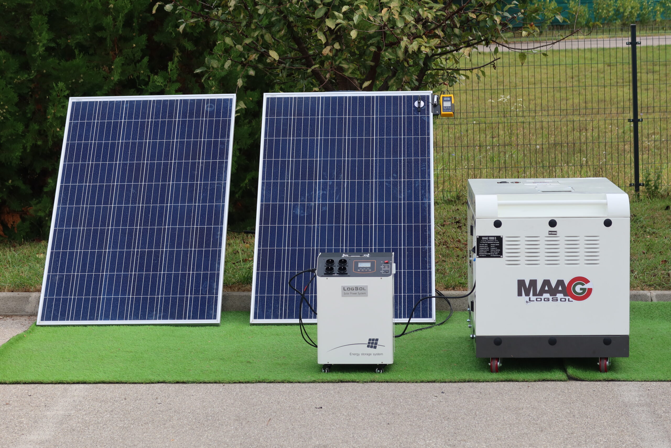 Két napelem, egy hordozható energiatároló egység és egy MAAG LOGSOL aggregátor látható szabadtéri bemutatóhelyen, zöld háttér előtt.
