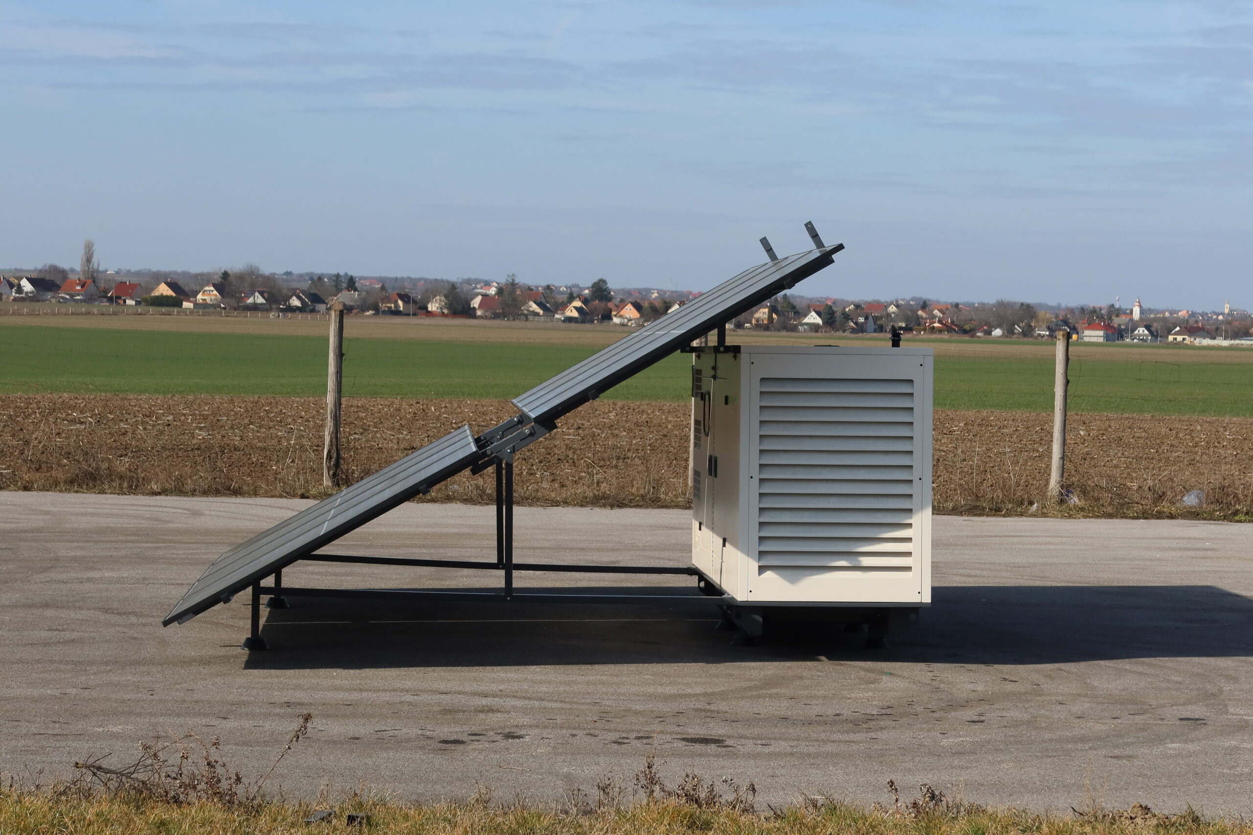 Oldalnézetű mobil napelemes aggregátor Alt text: Oldalról fotózott mobil napelemes aggregátorrendszer látható, megdöntött panelekkel és fehér gépházzal, mezőgazdasági terület és település háttérrel.